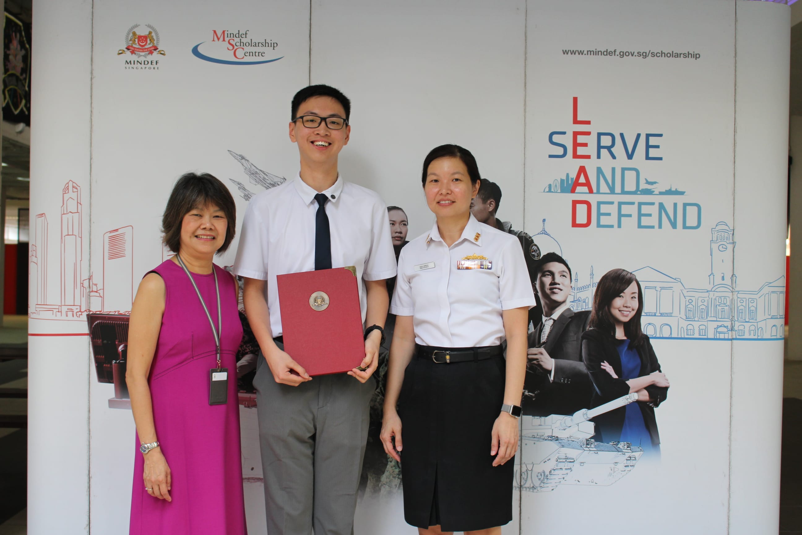 One student holding red folder stand with two adults, one in military uniform