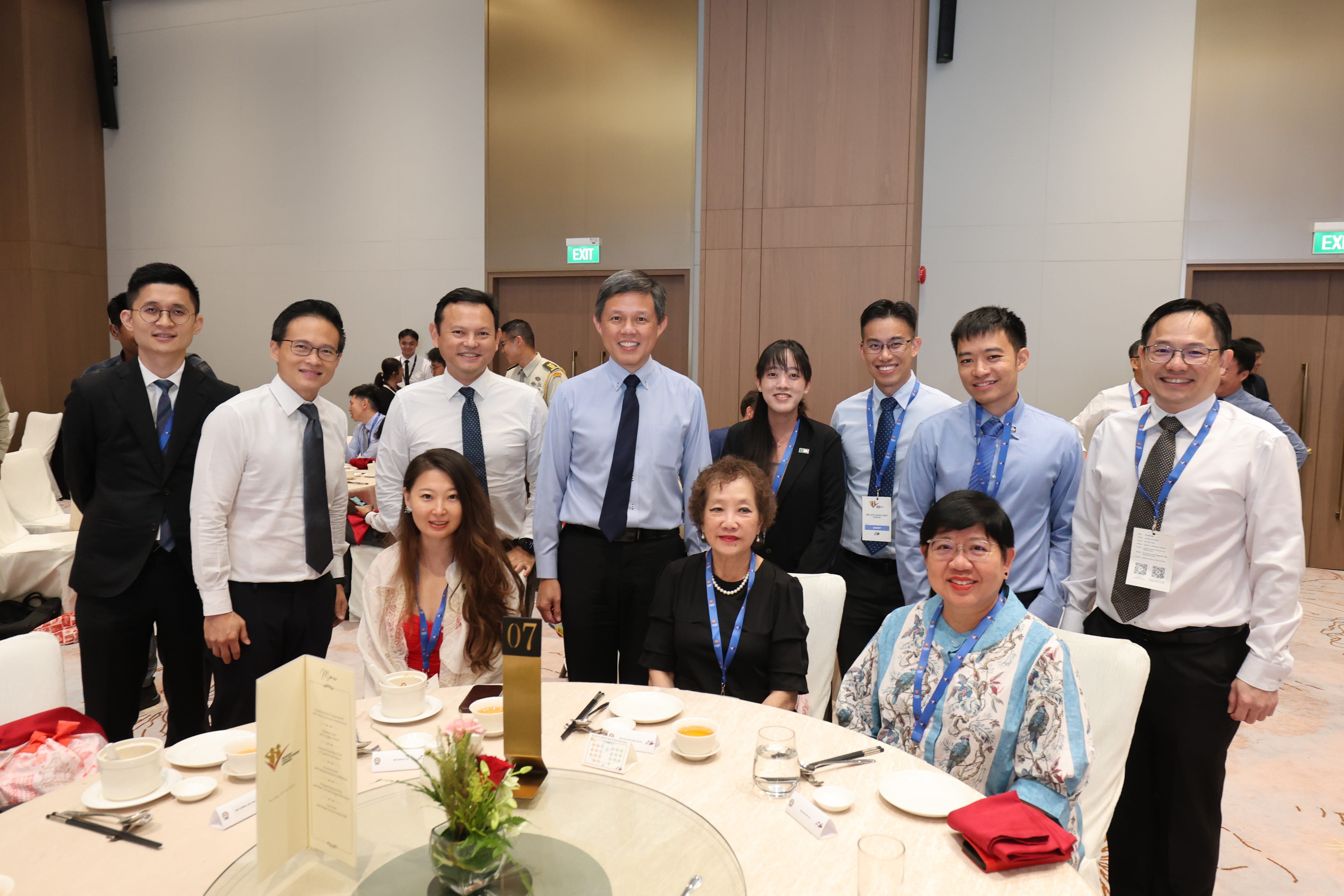 Group of people posing behind dining tables in a banquet hall with Exit signs visible.