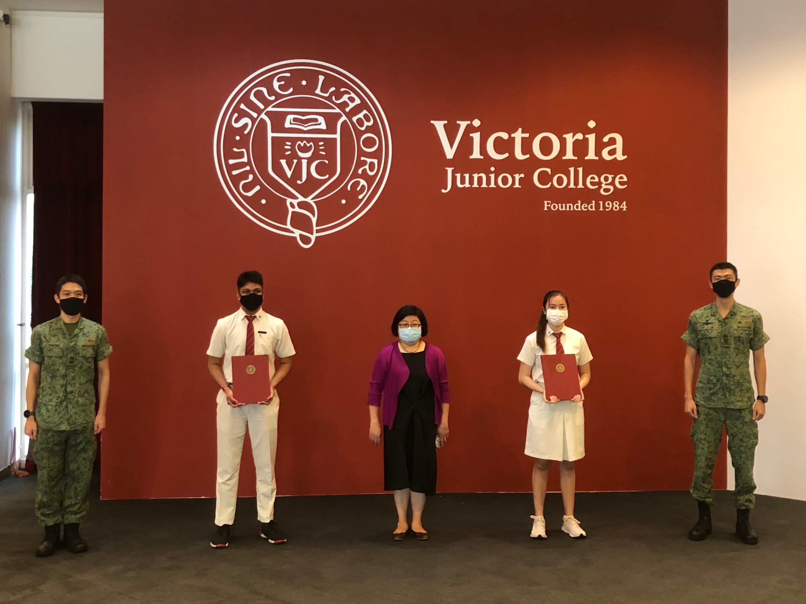Two students holding red folders stand with three adults, two in military uniforms