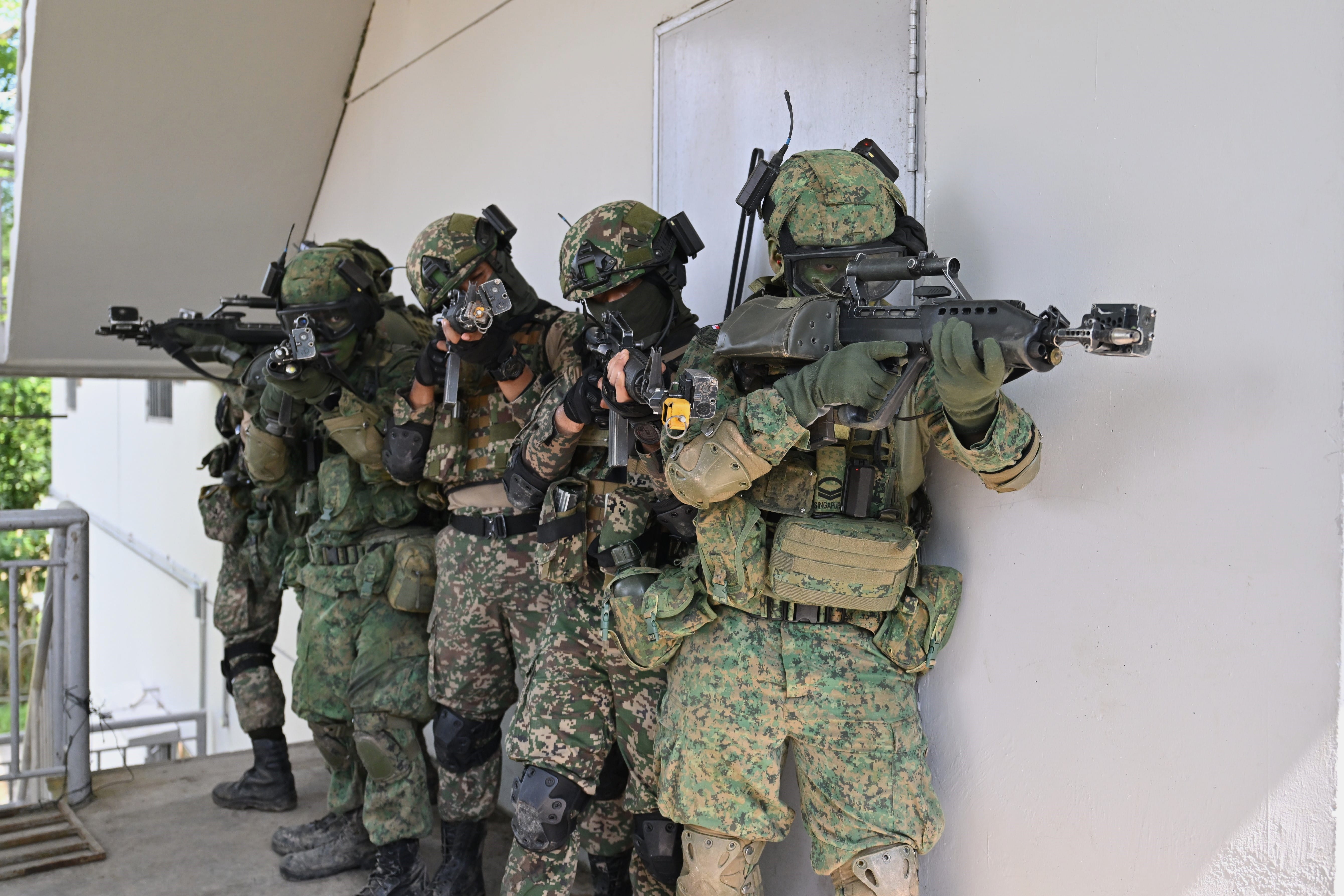 Soldiers in camouflage gear and helmets hold rifles in front of a white wall.