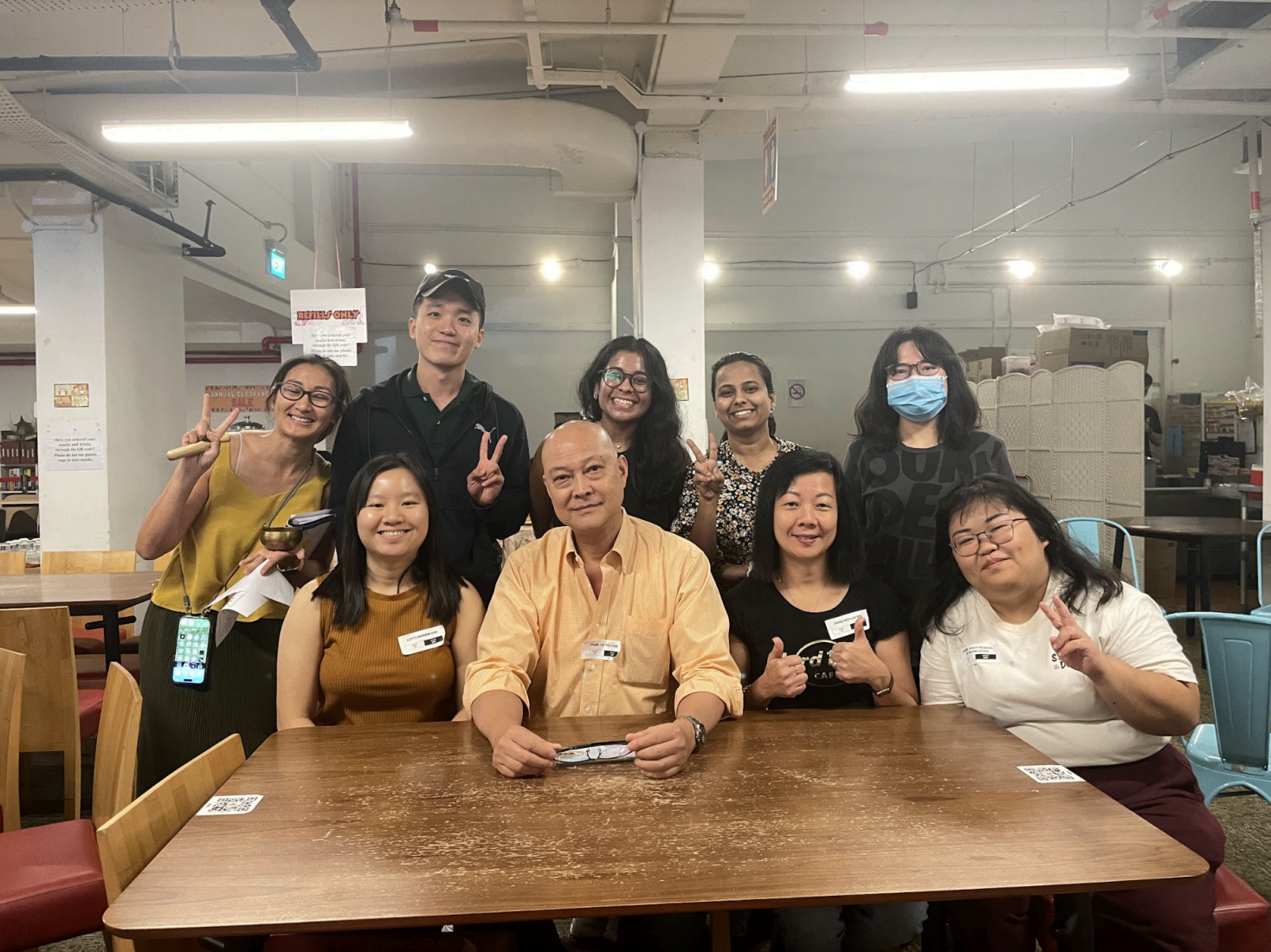 Nine smiling adults pose at a table, some giving peace signs, indoors.