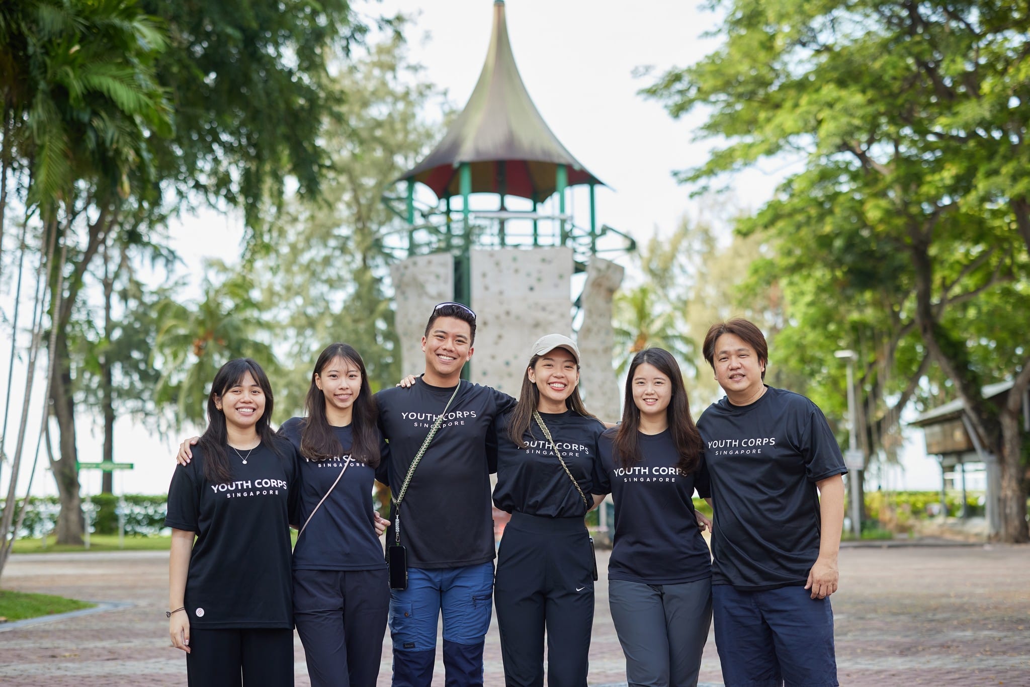 Six people in Youth Corps Singapore shirts pose in front of an outdoor climbing structure.
