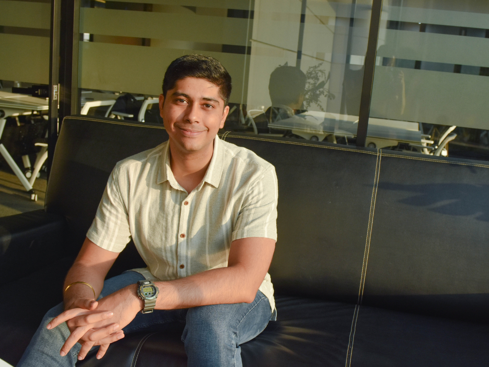 Dev Bahl seated on a black couch, smiling at the camera in a brightly lit room