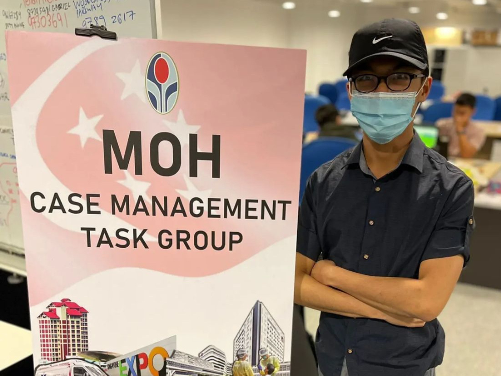 Toh Guan Ru in mask standing beside a MOH Case Management Task Group sign in an office