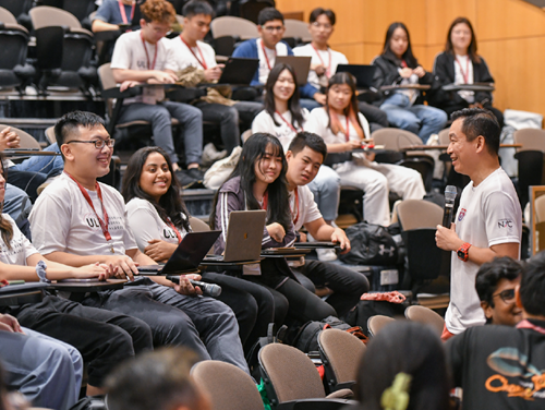 Lecture hall with Asian man speaking into mic to students with laptops