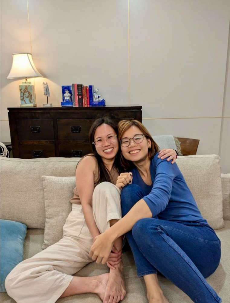 Two women with glasses sitting closely together on a beige couch, smiling.