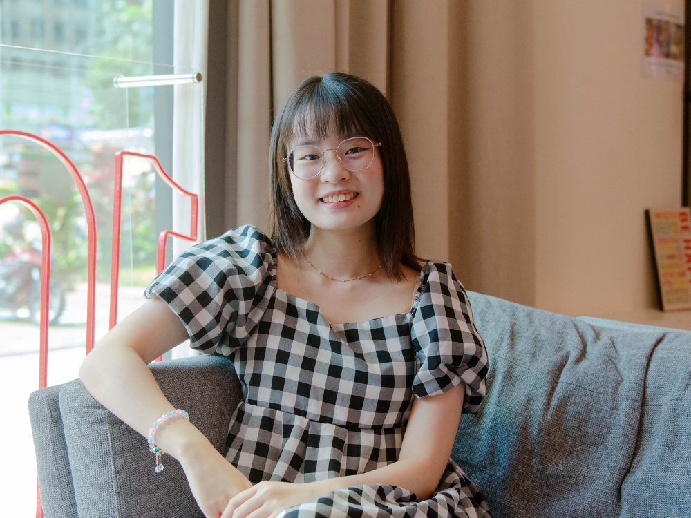 Erina in checkered dress smiling while seated on a sofa near a window