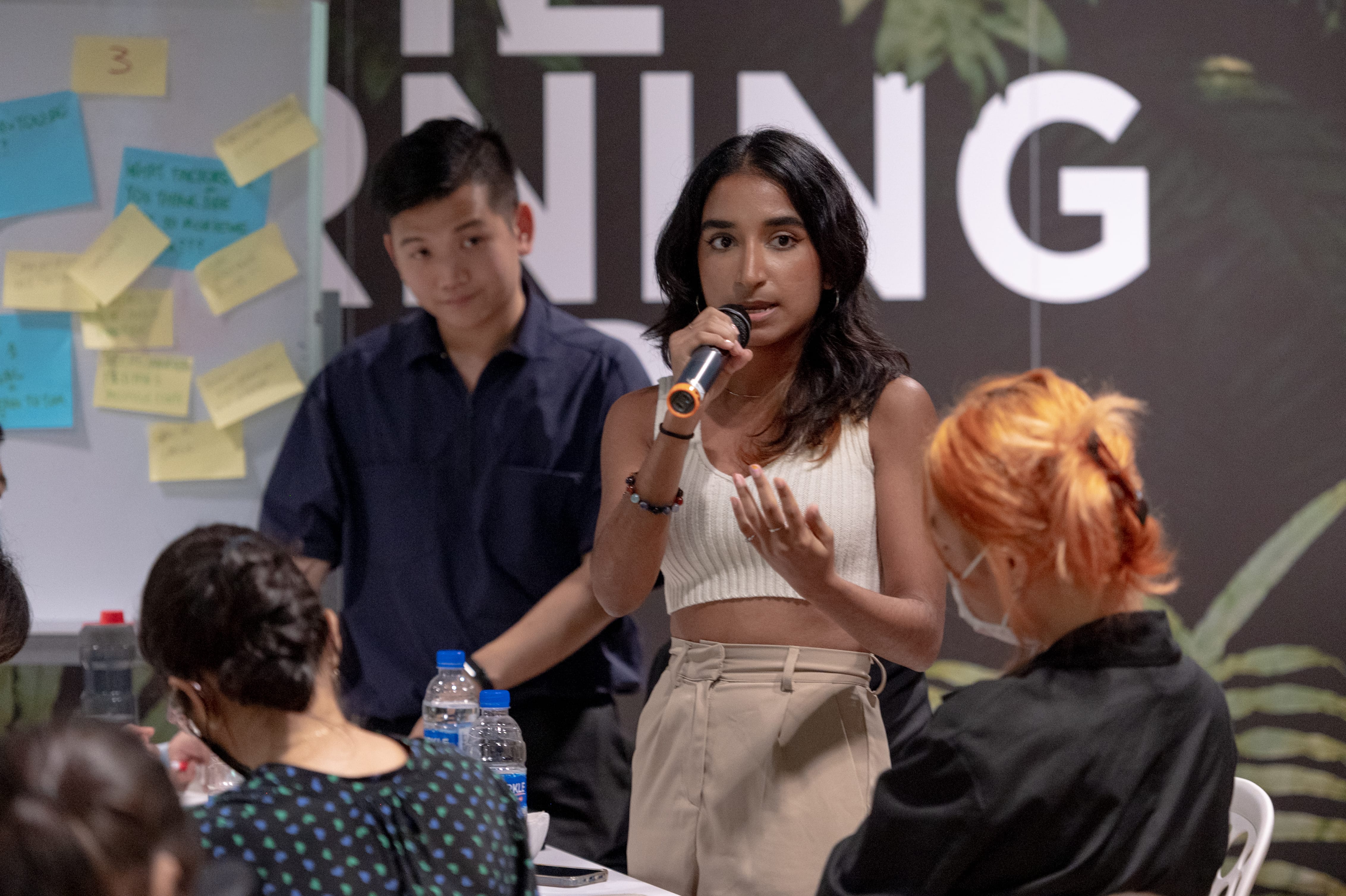 Person speaking into a microphone with audience members and a whiteboard behind.