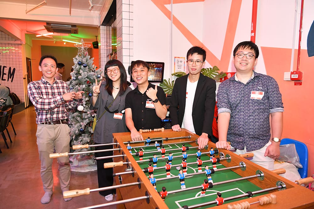 Four adults stand behind a foosball table in a decorated room.