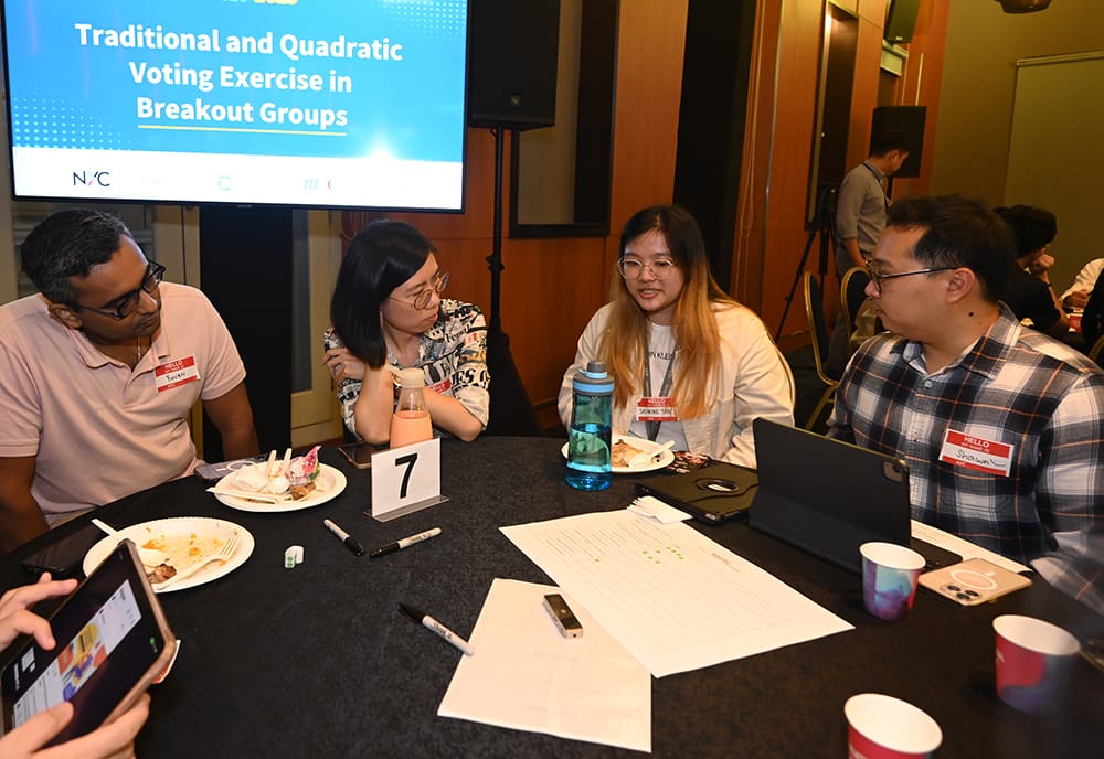Four people at table 7 with papers, tablets, and food under a presentation screen.