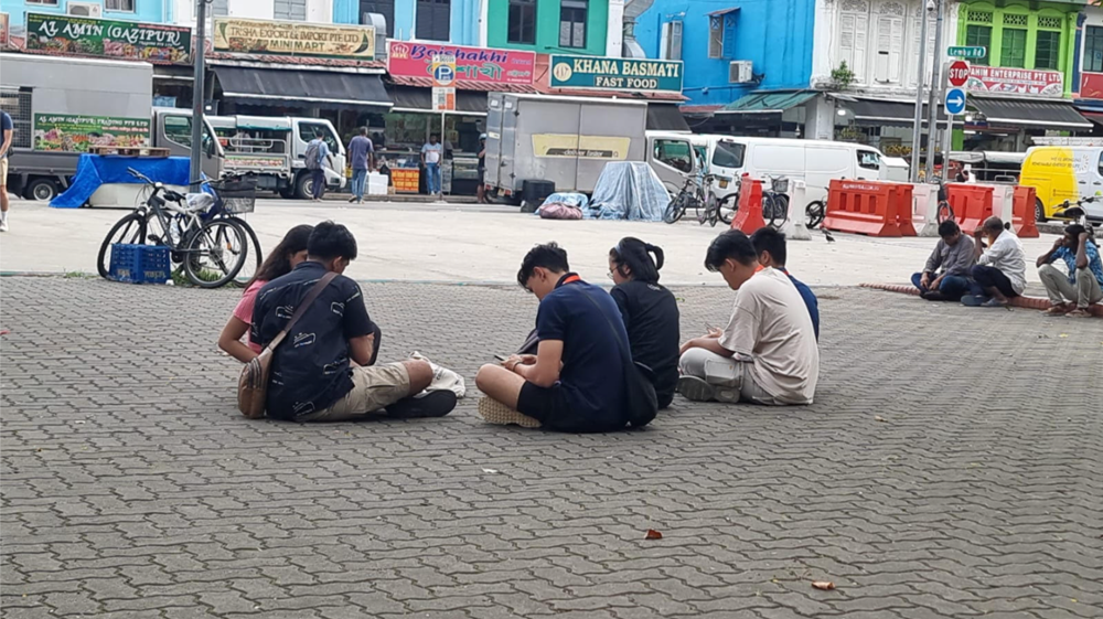 People sit on a brick plaza near storefronts and parked vehicles in a street setting.
