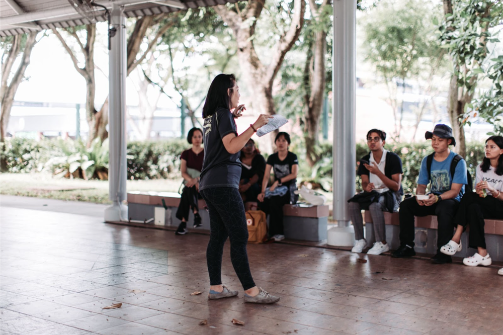 Woman presenting to a group seated outside on benches under a covered area.