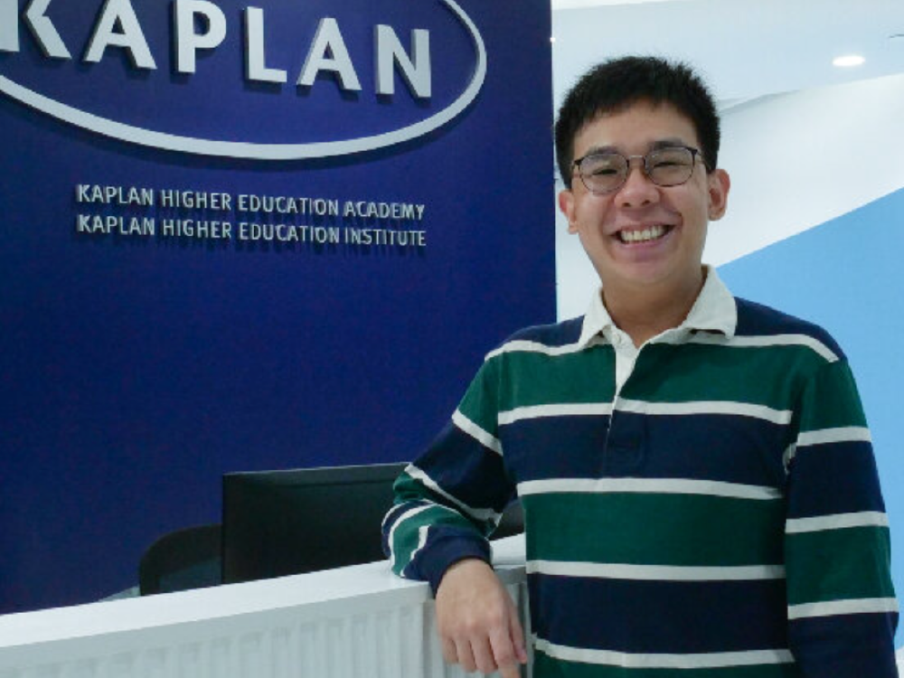 Julian stands in front of a Kaplan logo with a big smile