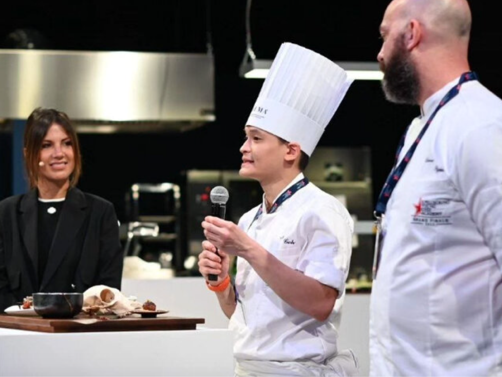 Chef Ian speaking into mic on stage with two others at a culinary presentation