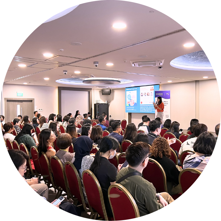 Conference room with speaker presenting to a seated audience, projector screen in view.