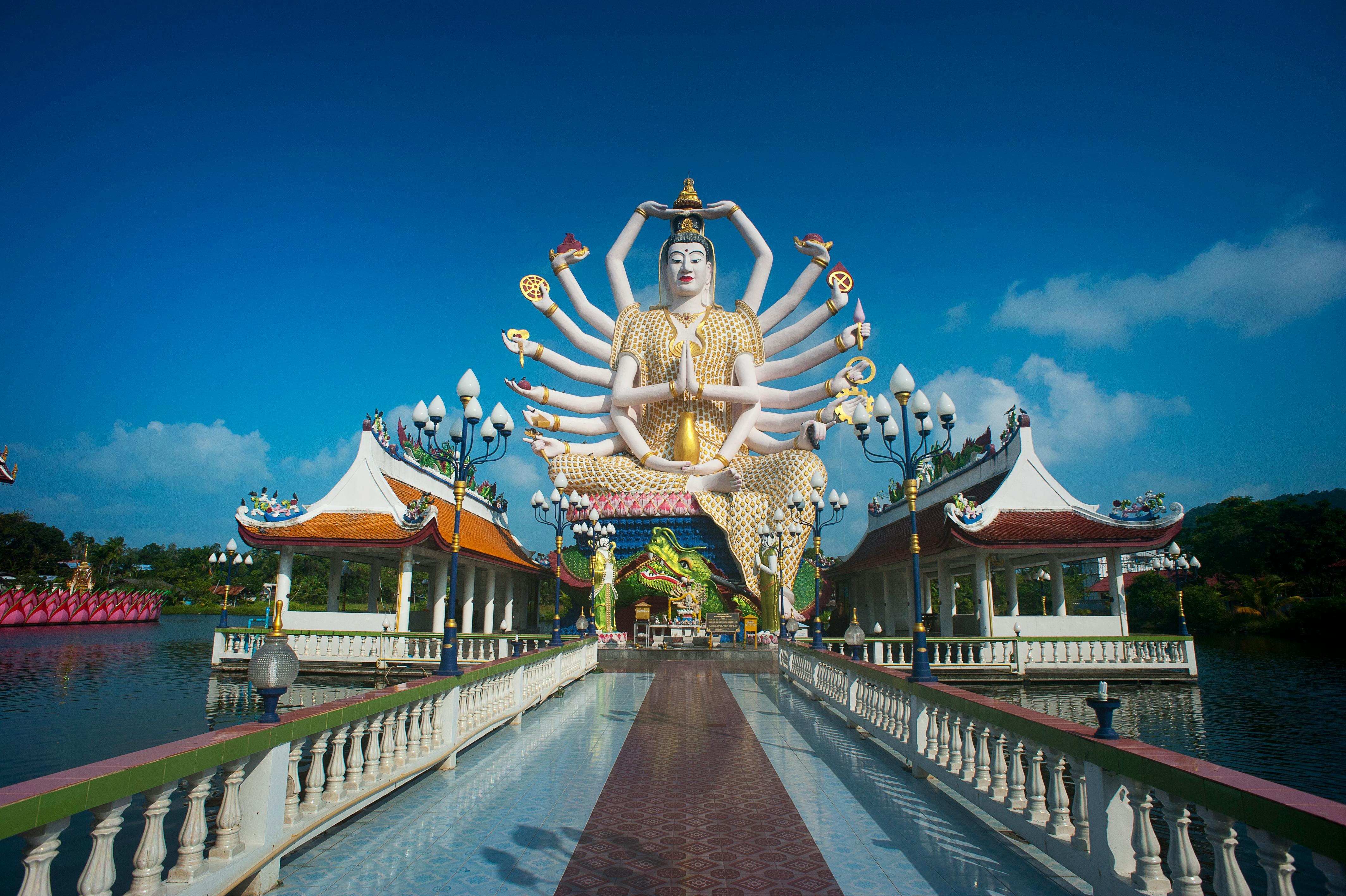Large, multi-armed white and gold Buddha statue in lotus pose, center of island temple. Blue sky.