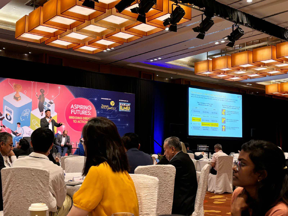 Conference room with speaker on stage, audience seated, and presentation screens.