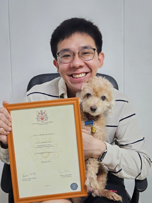 Julian holds a framed certificate while carrying a small dog and smiling