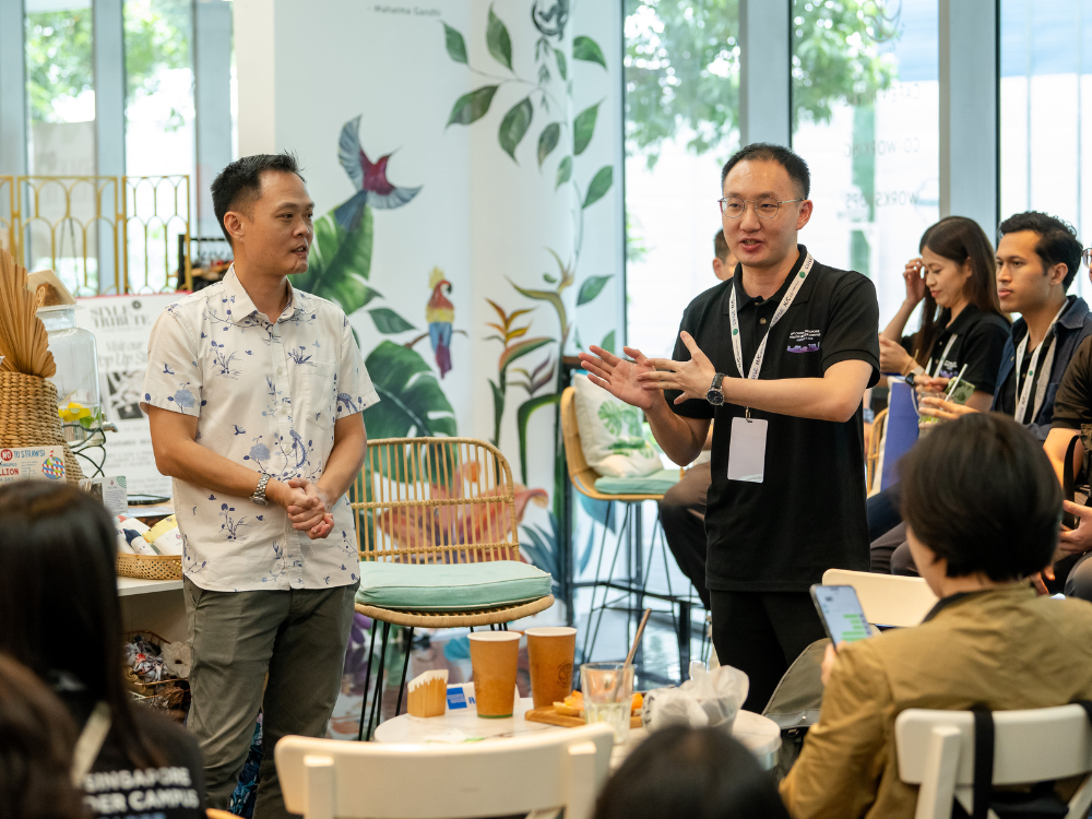 A man in black shirt with a lanyard speaking, beside a man in white shirt. Several participants sit around to listen.