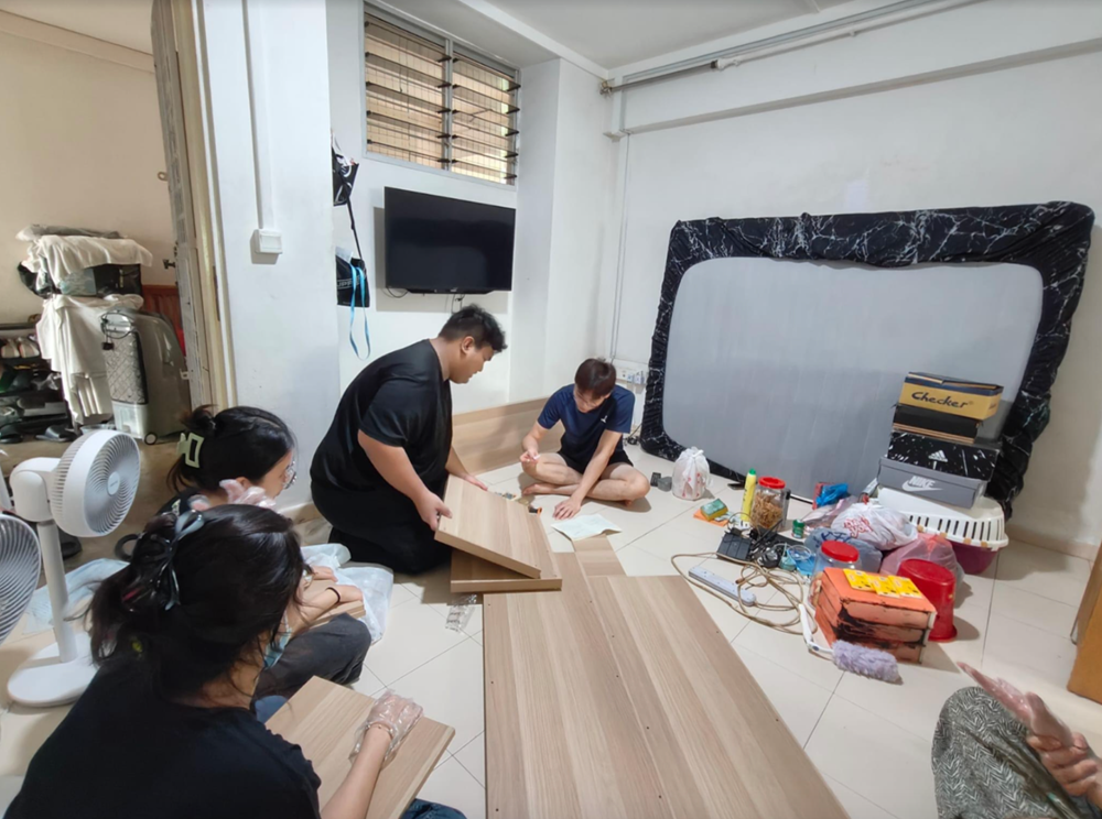 Four people assemble wood furniture on a tiled floor in a cluttered room.