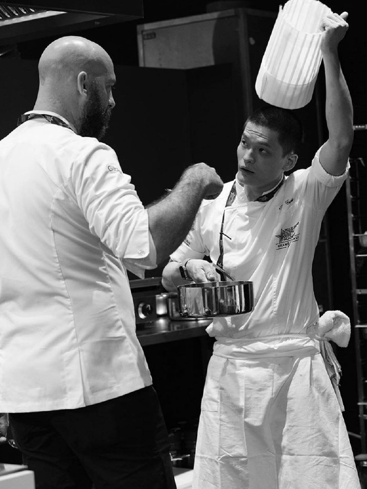 Chef Ian receiving feedback from mentor while holding a pot, in black and white