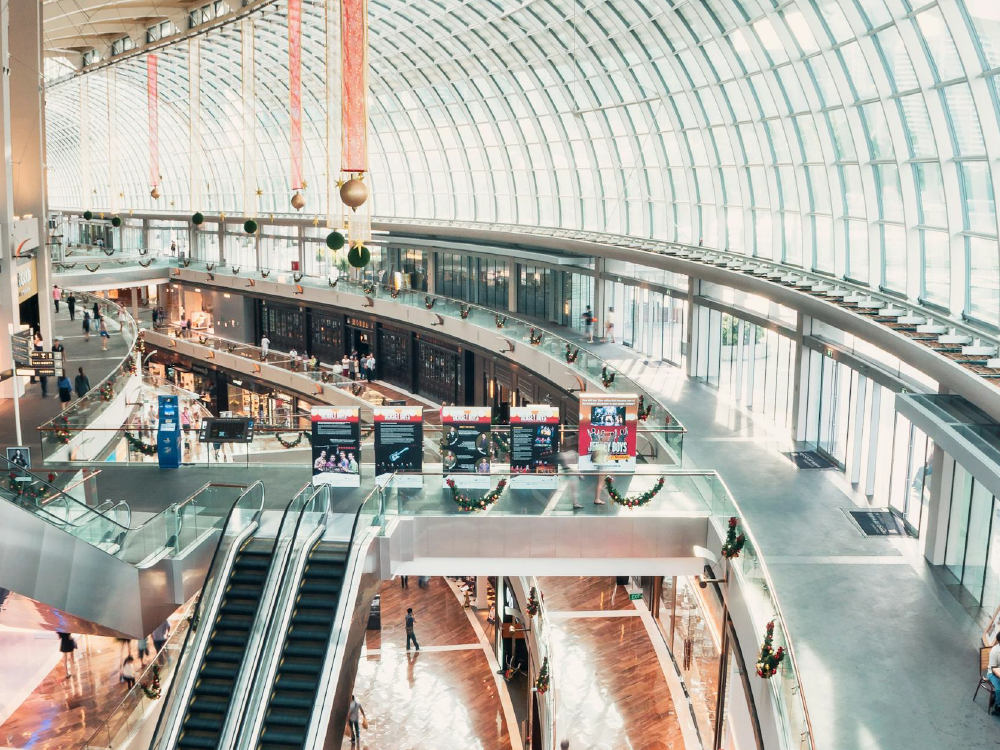 Multi-story shopping mall interior with glass roof, escalators, and people walking.