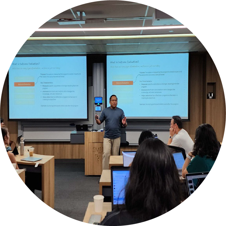 Classroom scene: Lecturer presents "Outcome Evaluation" on dual screens before seated students.
