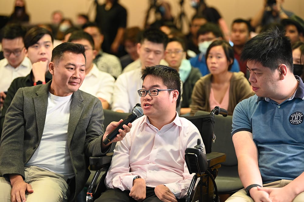 Person in wheelchair with microphone speaking to panel, audience visible.