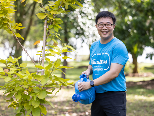 Mr Yen in a blue shirt smiling while watering a young tree at a green park