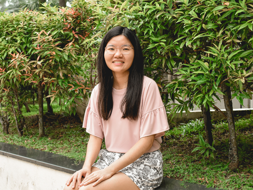 Anita smiling while seated outdoors near green shrubs, wearing a pink top