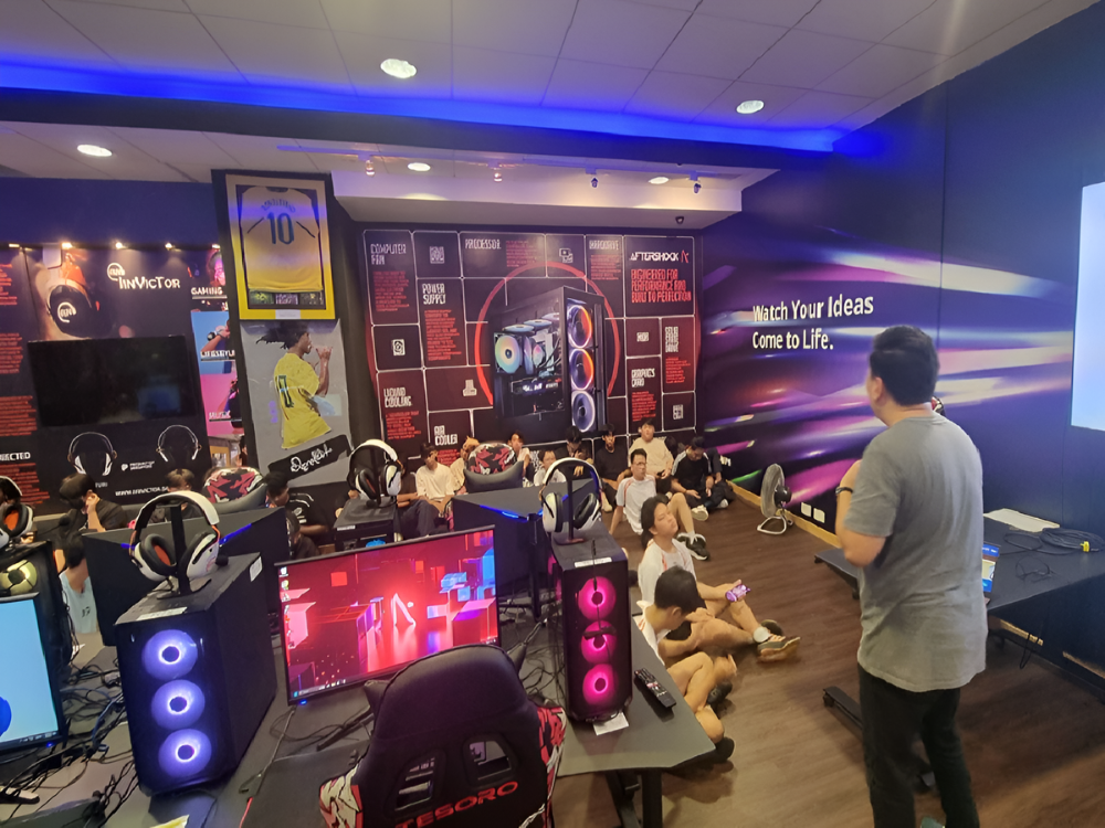 Inside gaming store, rows of PCs with headsets, wall art, person at front presenting.