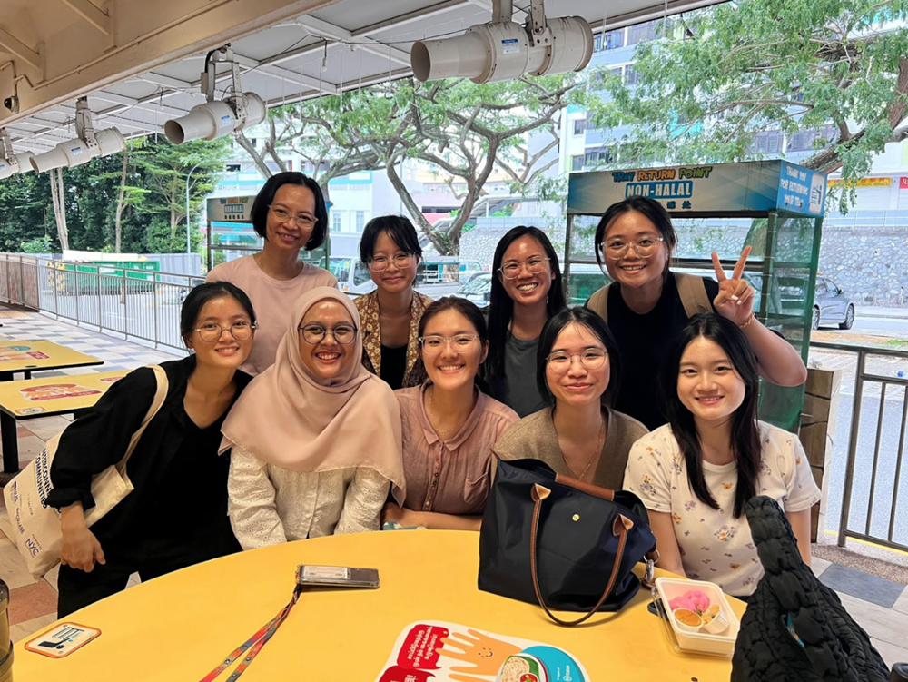 Group of nine smiling women with glasses pose around a yellow table outdoors.