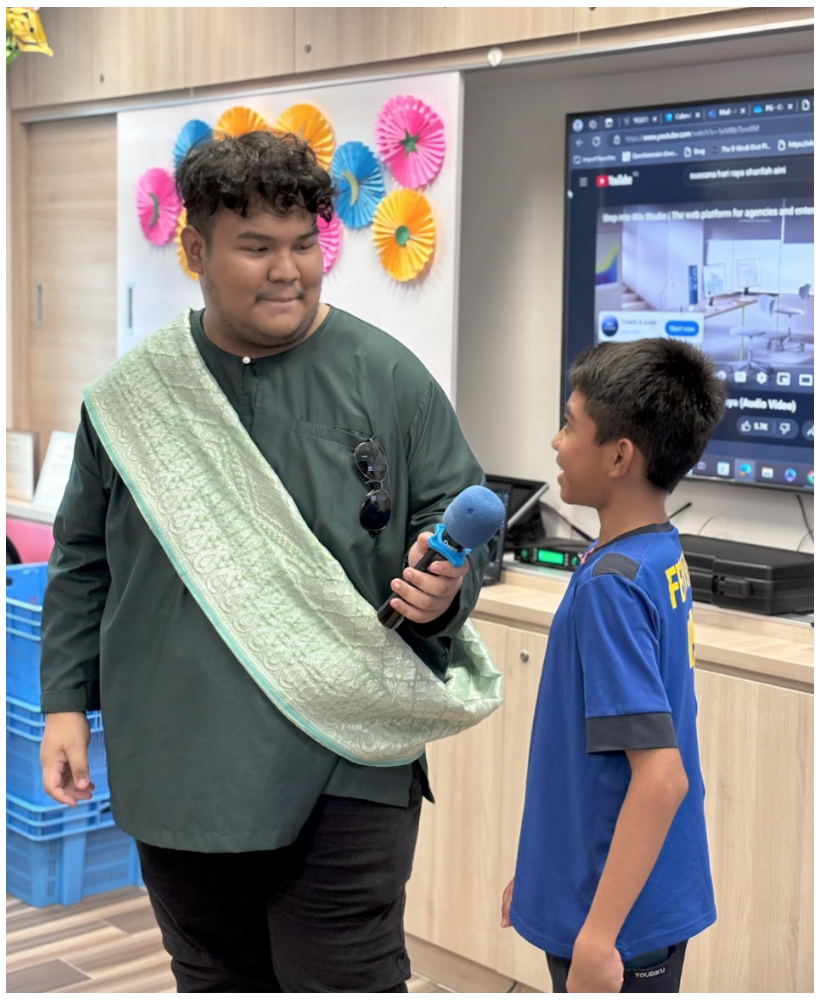 Man in green shirt, sash, holding mic talking to boy in blue jersey near TV screen.