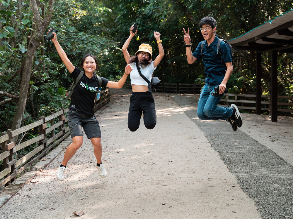 Three young adults jumping on a path in forest. One holds binoculars, another a phone.