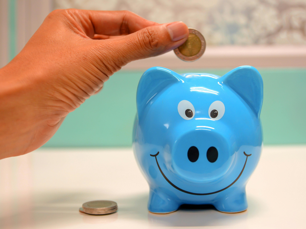 Hand dropping coin into a blue, smiling piggy bank. Another coin sits on the surface.