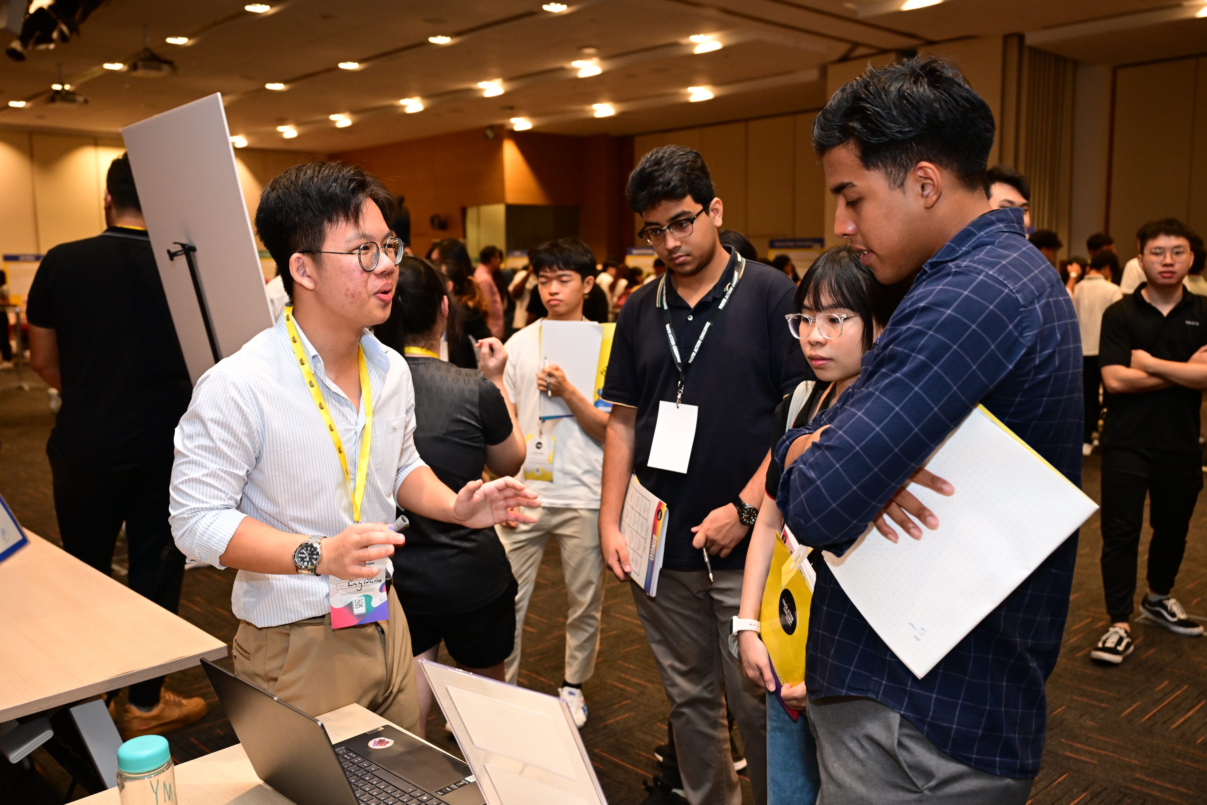 Group of young adults gathered around a table with a laptop, talking and holding notebooks.