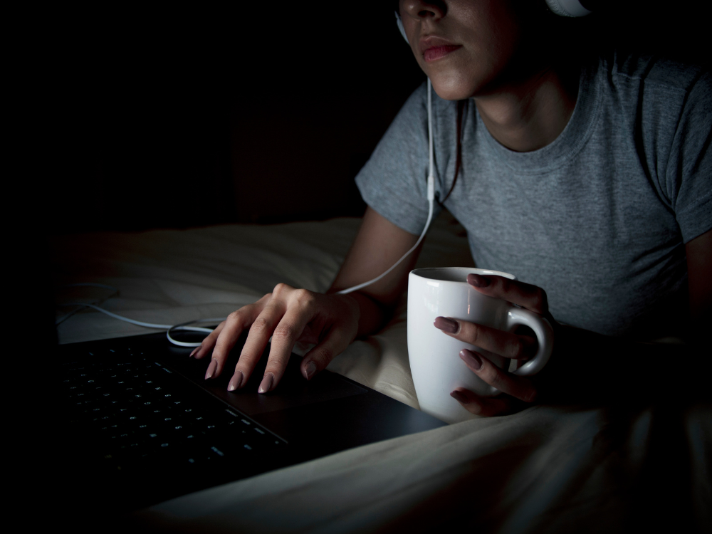 Person in bed with headphones, using laptop and holding a white mug in dim lighting.