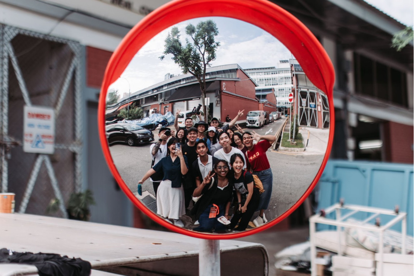 Group selfie mirrored in a red safety mirror on a street with cars and buildings.