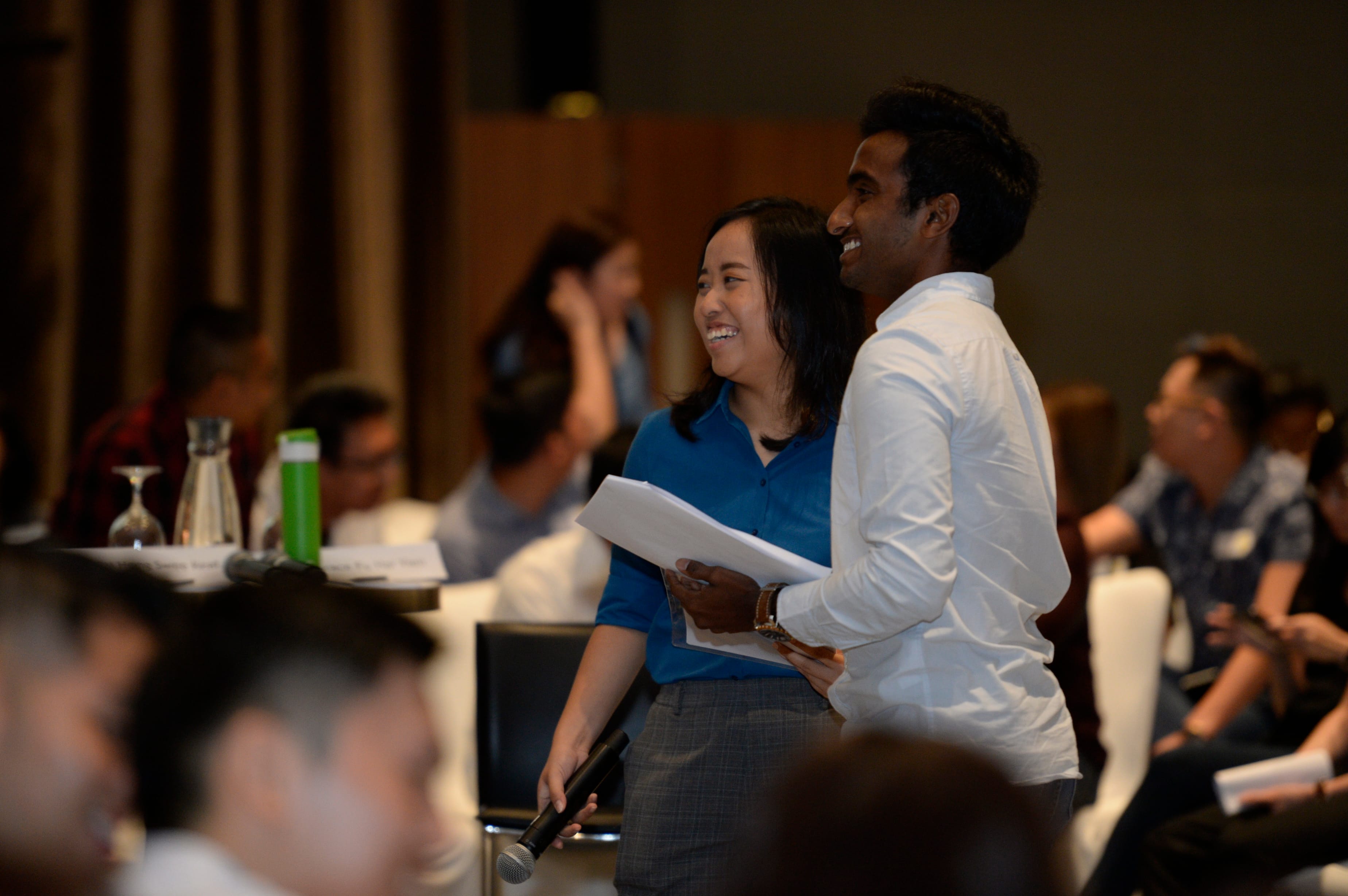 Two people in front of a crowd. Woman holding a microphone and papers, man in white shirt smiling.