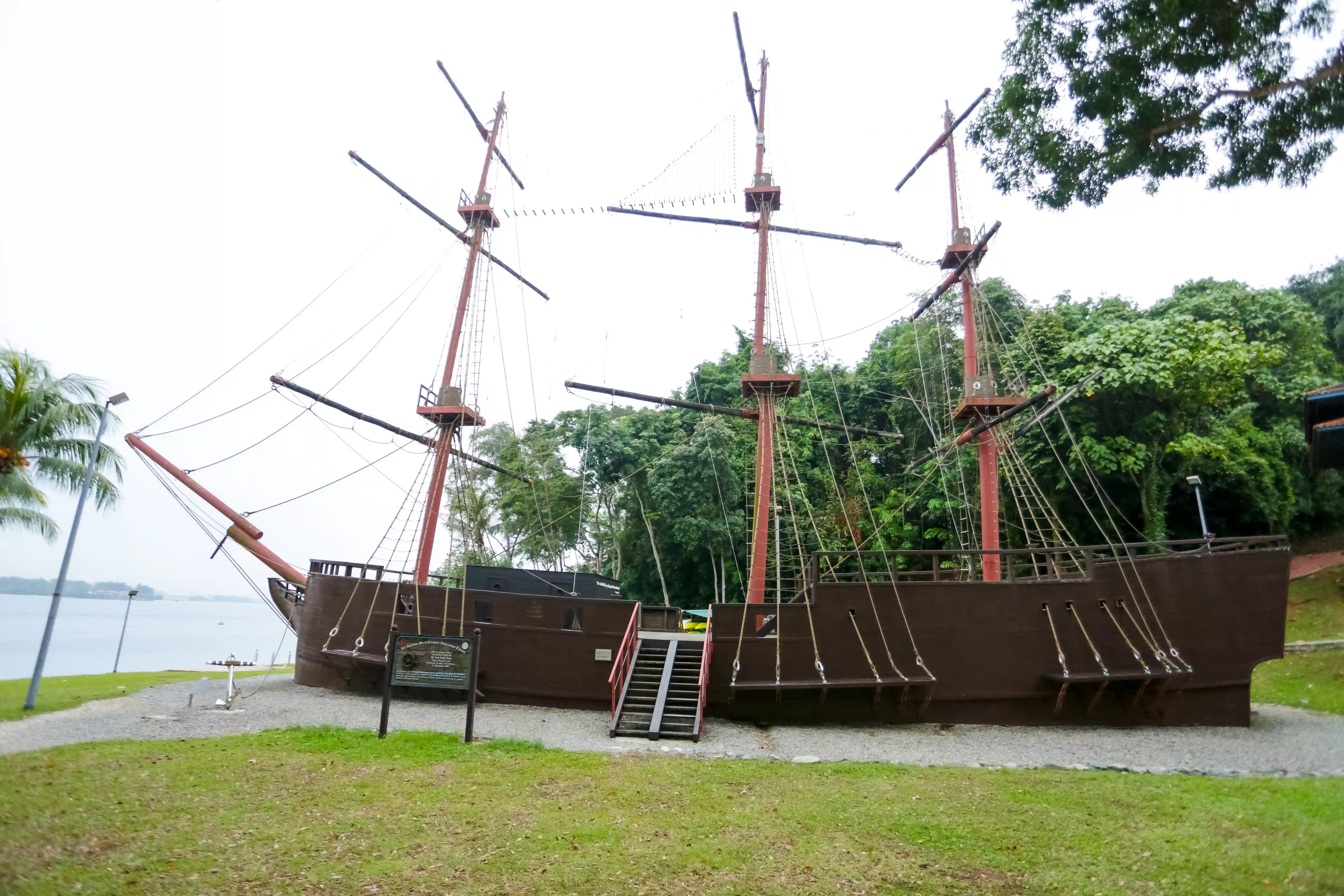 Large wooden replica ship with three masts and rigging positioned on grass near waterfront.