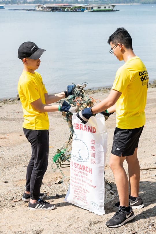 Two boys in yellow OBS shirts collect trash from a beach into a large sack.