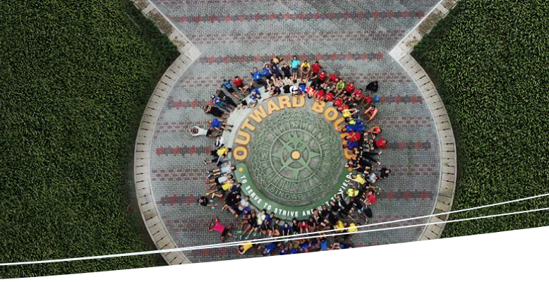 Aerial view of a diverse group of people forming a circle on a tiled plaza, surrounding the emblem of OBS.