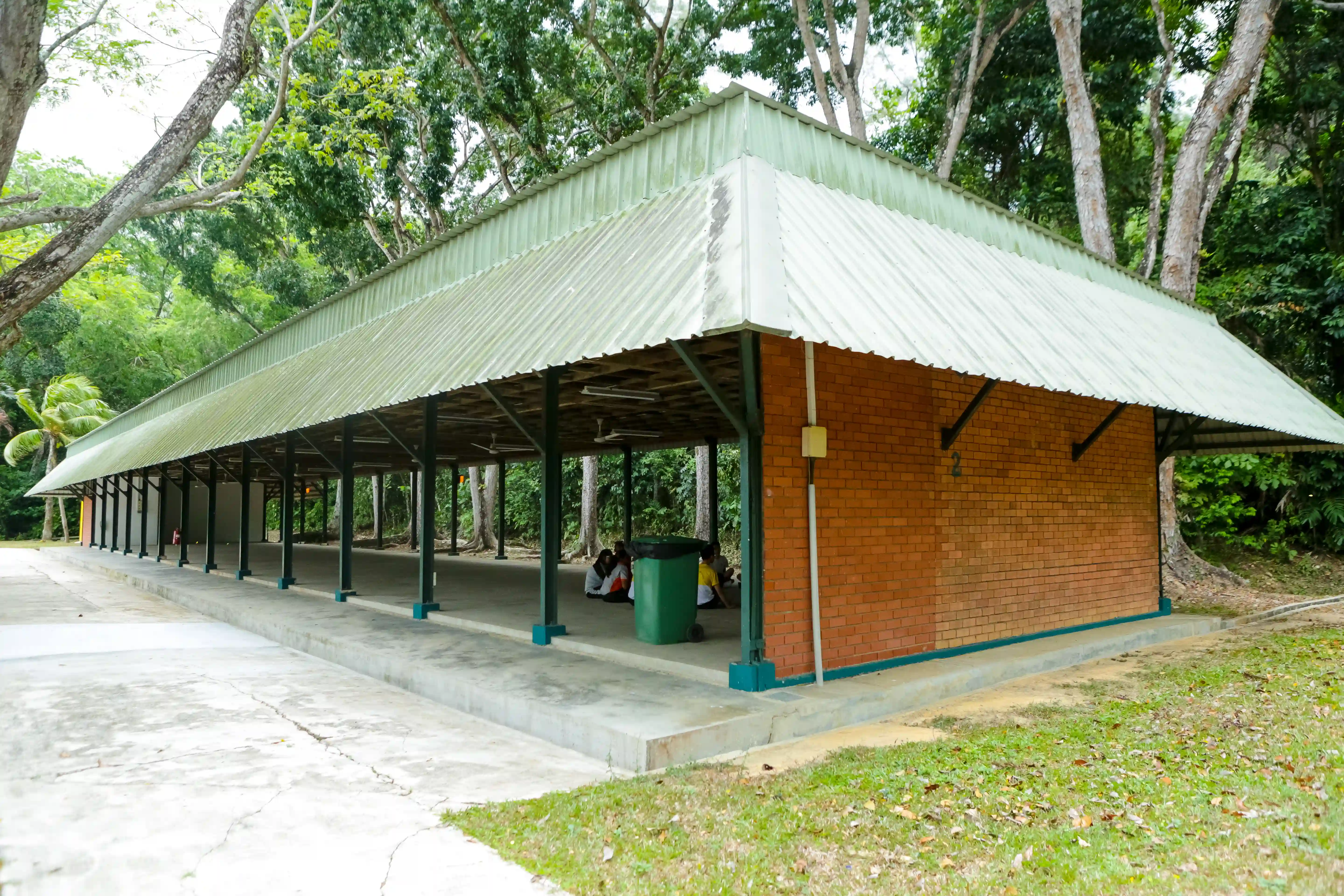 Open-air covered pavilion with green metal roof, brick walls, and concrete floor in forest.