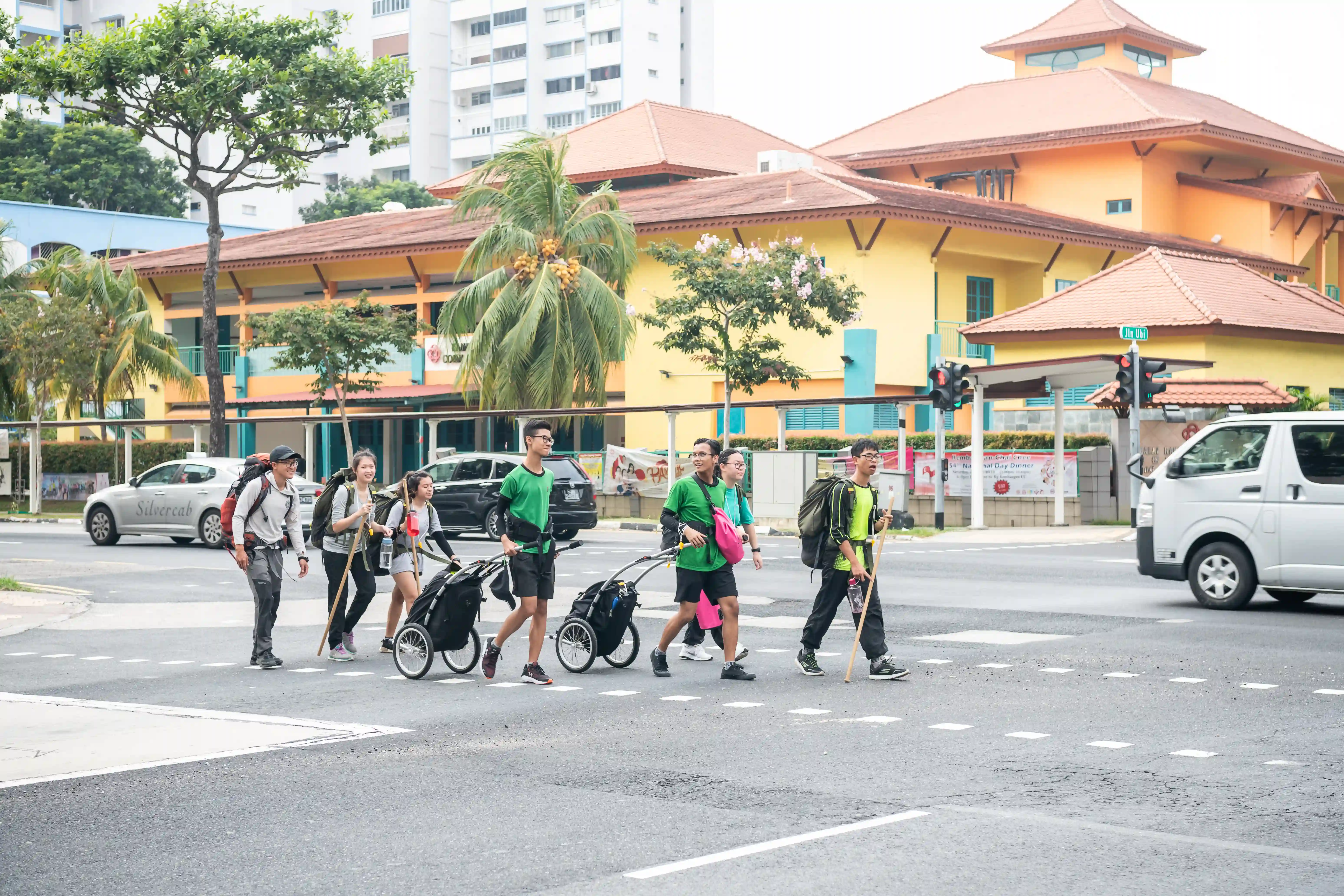 A group of people crossing a road with backpacks and walking sticks, some pulling carts with camping gear.