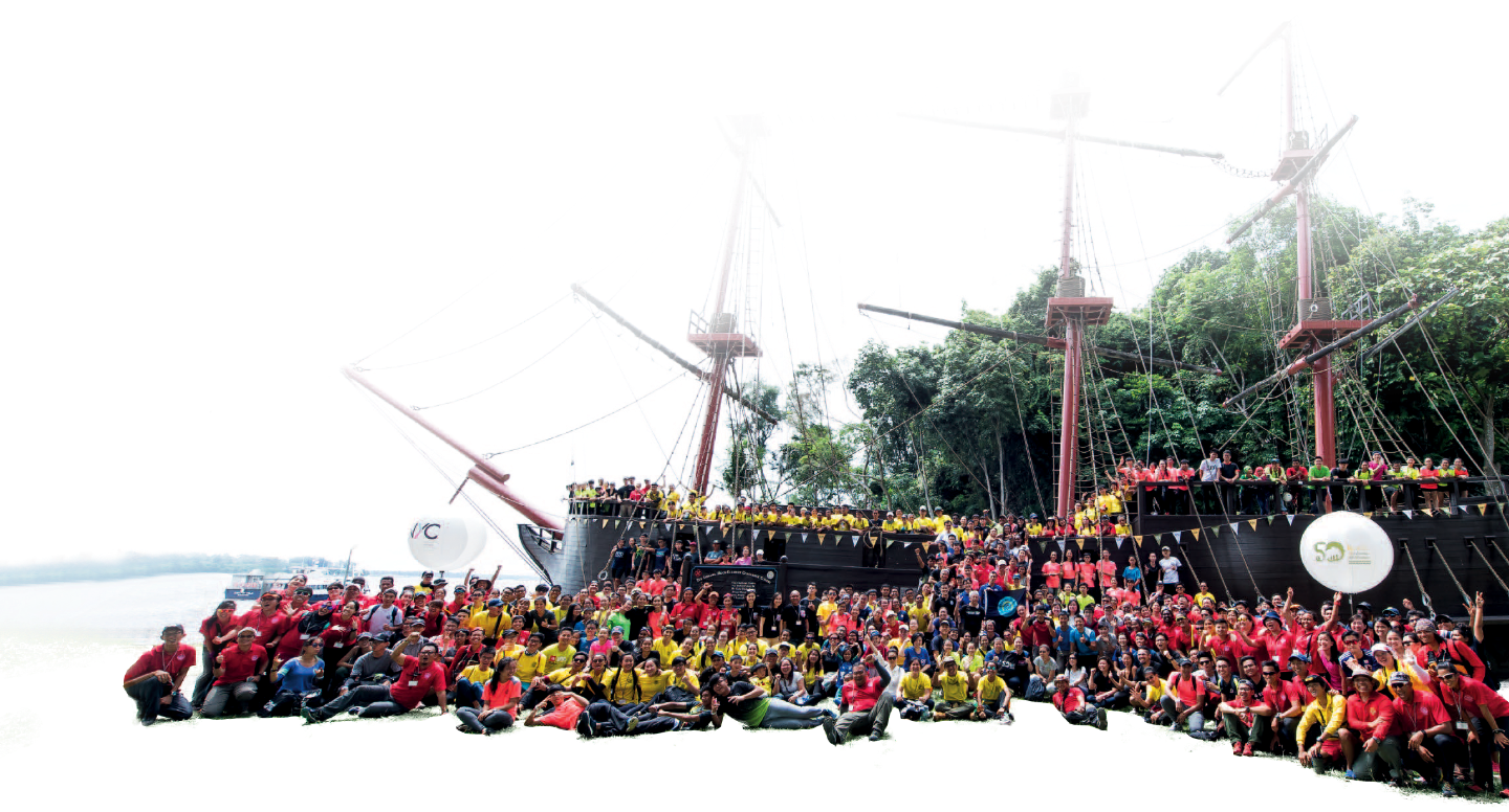 A large group of people posing for a group photo in front of a tall ship set against green trees.