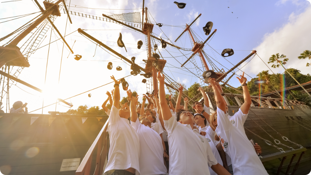 Youth in white shirts tossing caps beside a tall ship masts during OBS graduation.