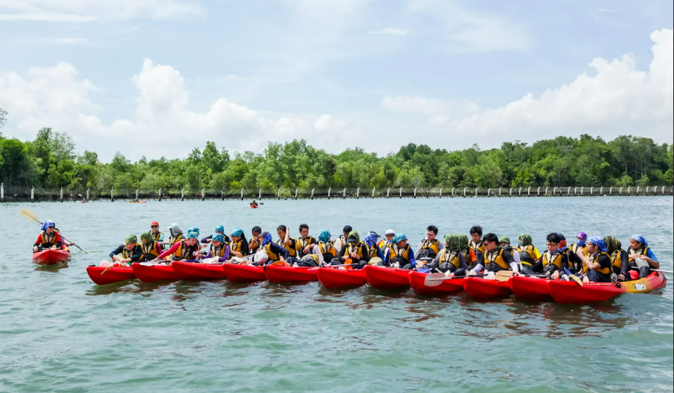 Large red kayak with many participants in life jackets paddling on calm water.