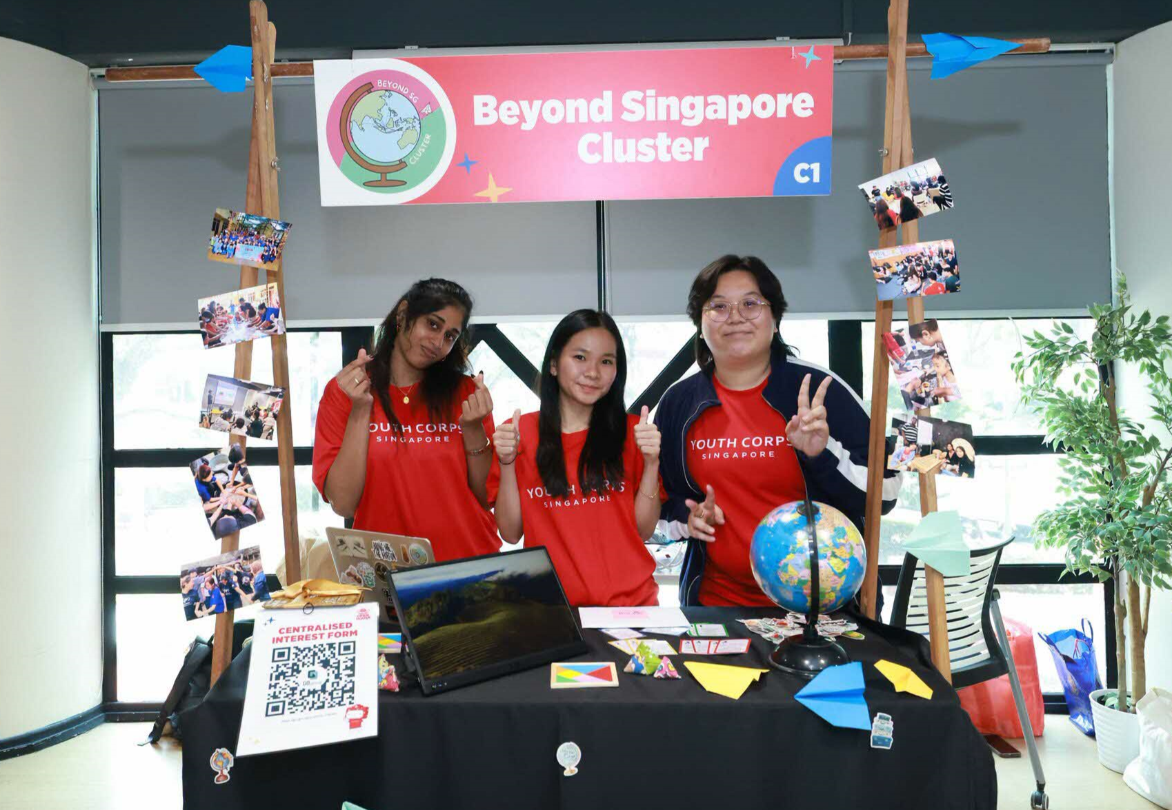 Youth Corps Singapore booth with volunteers, banner reading "Beyond Singapore Cluster", and flags displayed.