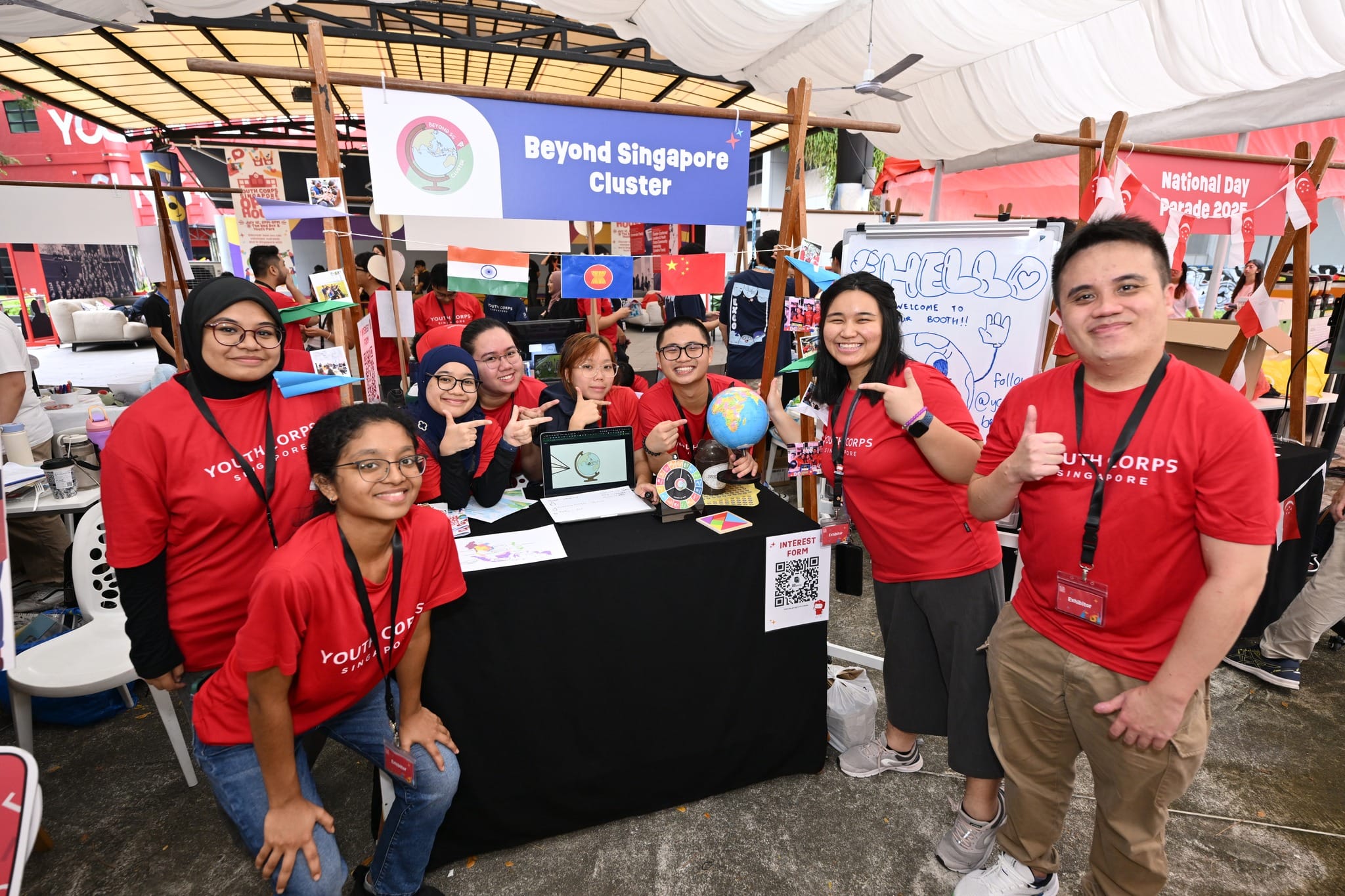 Youth Corps Singapore booth with volunteers, banner reading "Beyond Singapore Cluster", and flags displayed.