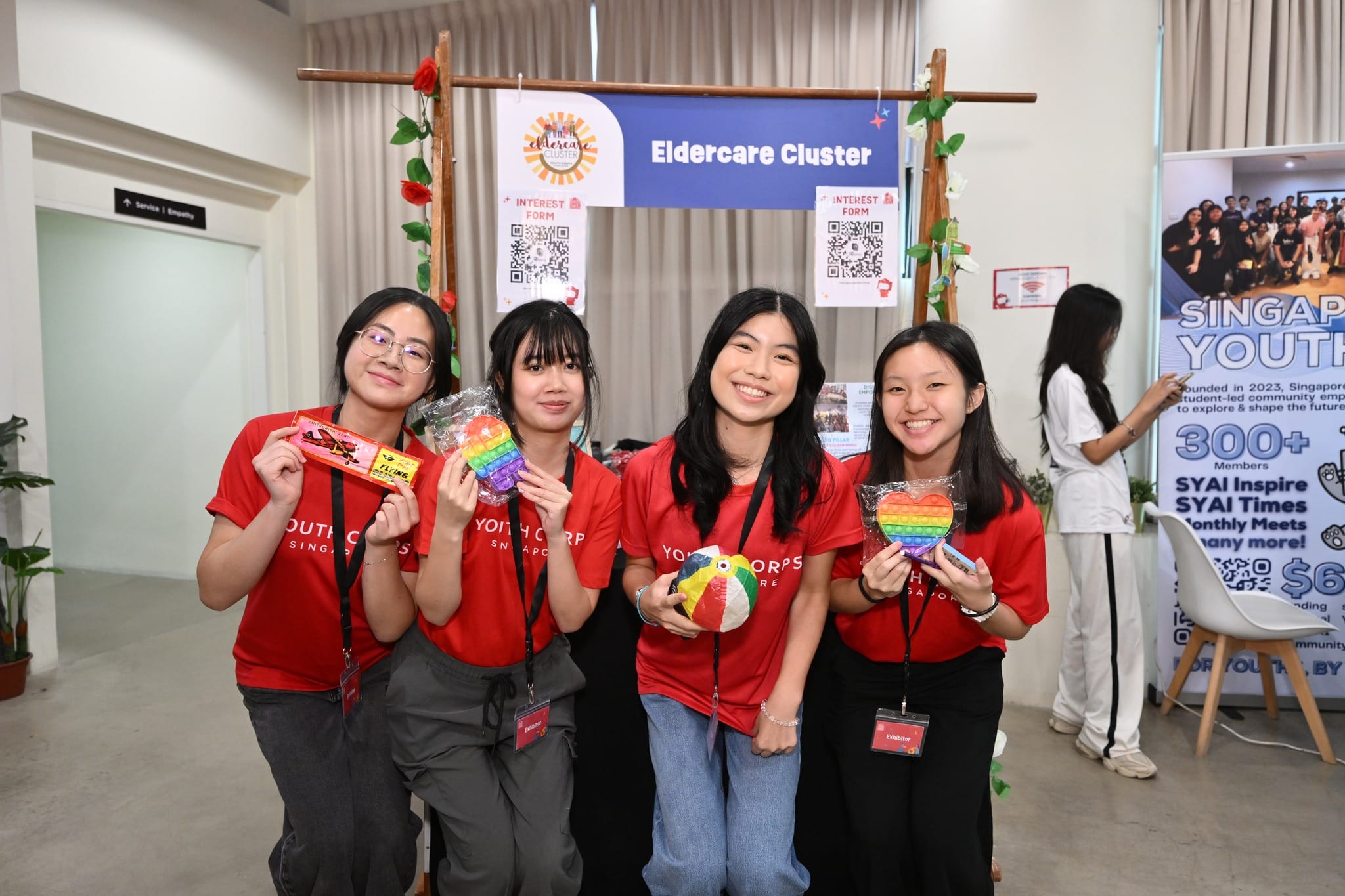 Four young women at an "Eldercare Cluster" booth holding toys, wearing red Youth Corps shirts.